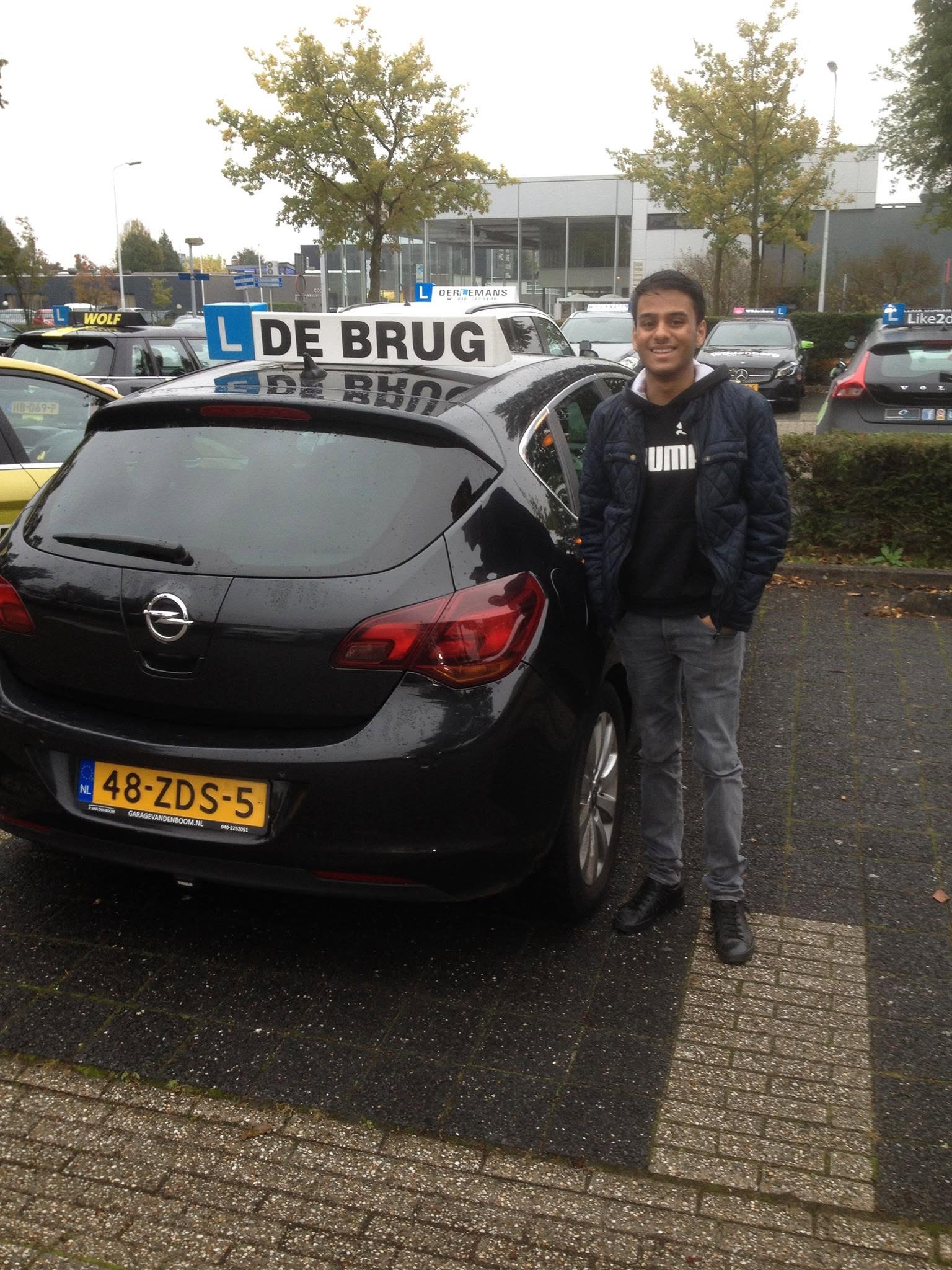 Geslaagde leerling naast de lesauto van Autorijschool De Brug op een regenachtige dag